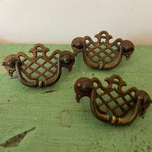 May include: Three antique brass drawer pulls with a decorative lattice design. The pulls have a curved handle and ornate detailing, perfect for vintage furniture restoration projects. The brass has a warm, aged patina.