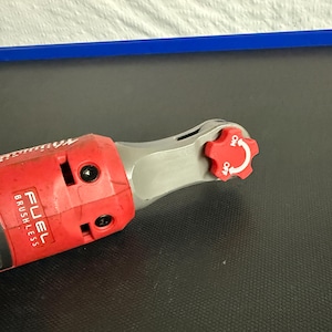 May include: A red and gray cordless ratchet wrench with the text "FUEL BRUSHLESS" visible. The wrench has a red, star-shaped control dial with "ON" and "OFF" markings. The tool is on a black surface.