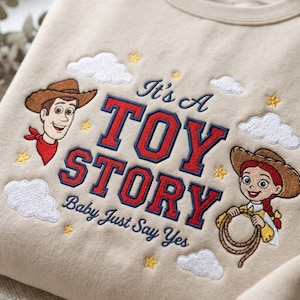 May include: A beige sweatshirt featuring embroidered characters Woody and Jessie from Toy Story. The design includes the text It's A TOY STORY Baby Just Say Yes in red and navy blue, surrounded by white clouds and small yellow stars.