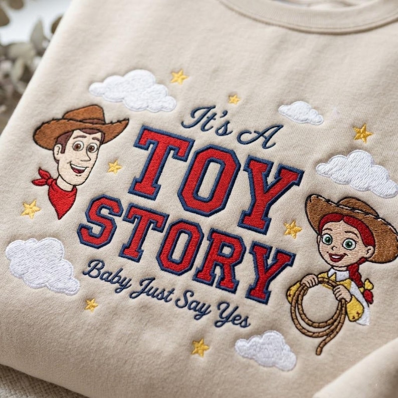 May include: A beige sweatshirt featuring embroidered characters Woody and Jessie from Toy Story. The design includes the text It's A TOY STORY Baby Just Say Yes in red and navy blue, surrounded by white clouds and small yellow stars.