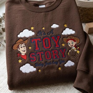May include: A brown sweatshirt featuring embroidered characters Woody and Jessie from Toy Story. The design includes the text "It's A TOY STORY" and "Baby Just Say Yes" in red and blue, surrounded by white clouds and yellow stars.