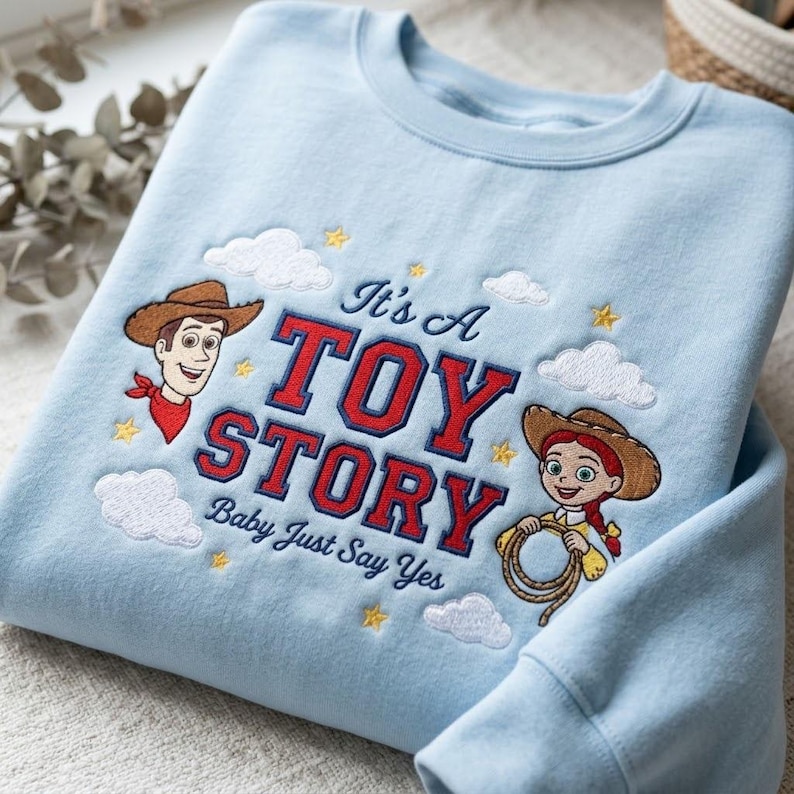 May include: A light blue crewneck sweatshirt featuring embroidered characters Woody and Jessie from Toy Story. The design includes red and blue text reading It's A TOY STORY Baby Just Say Yes, surrounded by white clouds and yellow stars.