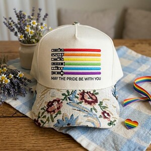 Puede incluir: Gorra de b&eacute;isbol crema con visera estampada floral. La gorra presenta un dise&ntilde;o de arco&iacute;ris con el texto "MAY THE PRIDE BE WITH YOU". El dise&ntilde;o incluye colores rojo, naranja, amarillo, verde, azul y morado.