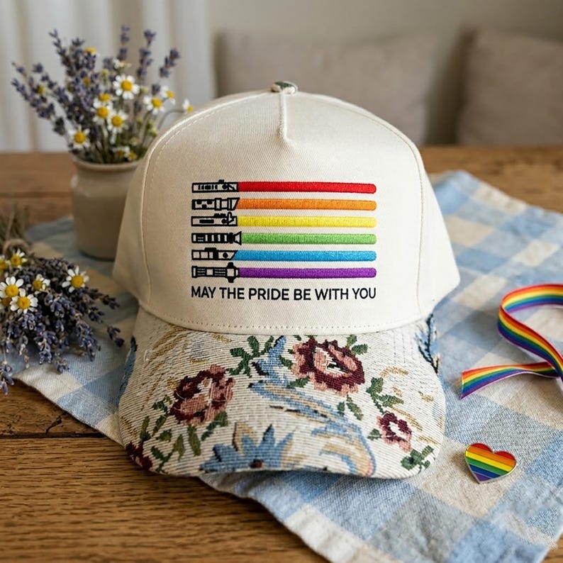 Puede incluir: Gorra de b&eacute;isbol crema con visera estampada floral. La gorra presenta un dise&ntilde;o de arco&iacute;ris con el texto "MAY THE PRIDE BE WITH YOU". El dise&ntilde;o incluye colores rojo, naranja, amarillo, verde, azul y morado.