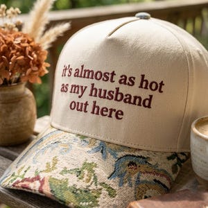 May include: A beige baseball cap with the embroidered text "it's almost as hot as my husband out here." The brim features a floral tapestry design in shades of blue, green, and pink. A small, round, gray button is on top of the cap.