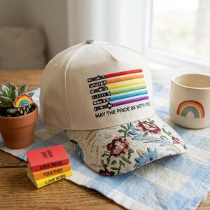 Puede incluir: Gorra de b&eacute;isbol beige con visera estampada floral y dise&ntilde;o de sable de luz arco&iacute;ris. El texto "MAY THE PRIDE BE WITH YOU" est&aacute; bordado debajo. Tambi&eacute;n se ve un peque&ntilde;o pin de arco&iacute;ris y una taza con un arco&iacute;ris.