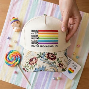 Puede incluir: Gorra de b&eacute;isbol color crema con un dise&ntilde;o de sable de luz arco&iacute;ris y el texto "MAY THE PRIDE BE WITH YOU". La gorra tiene una visera con estampado floral. Tambi&eacute;n se ven una piruleta arco&iacute;ris y una tarjeta "Happy Pride".
