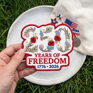 May include: A white embroidered patch with a red border featuring the number 250 decorated with green leaves, white daisies, red poppies, and blue flowers. Below, the text reads YEARS OF FREEDOM and 1776 - 2026 in red and blue embroidery.