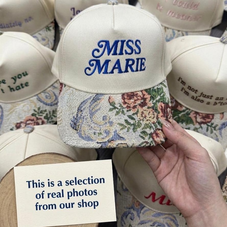 Puede incluir: Gorra de b&eacute;isbol color crema con "MISS MARIE" bordado en azul. La visera presenta un dise&ntilde;o de tapiz floral con tonos rosas, marrones y azules. Se ven otras gorras con texto bordado.