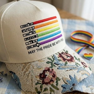 Puede incluir: Gorra de b&eacute;isbol color crema con un dise&ntilde;o de sable de luz arco&iacute;ris y el texto "MAY THE PRIDE BE WITH YOU". La visera presenta un estampado floral. Una cinta arco&iacute;ris y un pin en forma de coraz&oacute;n son visibles.