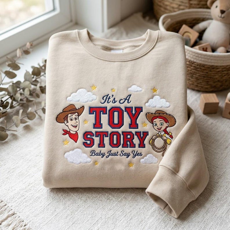 May include: A beige crewneck sweatshirt featuring embroidered characters Woody and Jessie from Toy Story. The design includes the text "It's A TOY STORY Baby Just Say Yes" in red and blue, surrounded by embroidered white clouds and yellow stars.