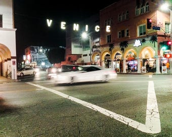 Peut inclure: Vue nocturne d'une rue à Venice, en Californie. La rue est bordée de bâtiments et de palmiers. Le panneau "VENICE" est éclairé en lettres blanches. Une voiture blanche roule dans la rue.