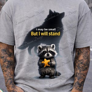 May include: Gray t-shirt featuring a raccoon holding a yellow star, with a black wolf silhouette and the text "I may be small But I will stand." The design is printed on a short-sleeved shirt.