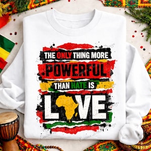 May include: A white long-sleeve sweatshirt featuring a graphic with red, yellow, and green distressed brushstroke backgrounds. The text reads: THE ONLY THING MORE POWERFUL THAN HATE IS LOVE, with the letter O replaced by a yellow map of Africa.