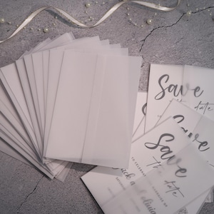 May include: A selection of white translucent envelopes and "Save the Date" cards. The cards display elegant calligraphy with the text "Save the date" and "To celebrate the marriage of Jack and Olivia". A ribbon and pearls are in the background.
