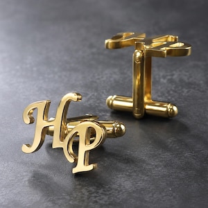 Op de afbeelding: Een paar goudkleurige metalen manchetknopen met de gestileerde letters H en P in een cursief lettertype. Elke manchetknop heeft een gepolijste afwerking en een standaard T-vormig draaimechanisme aan de achterkant van het monogramontwerp.