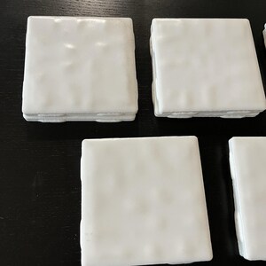 May include: A set of square, white ceramic coasters. Each coaster has a slightly textured surface and a raised edge. The coasters are arranged on a dark surface, showcasing their clean, minimalist design. Ideal for protecting surfaces from drink rings.