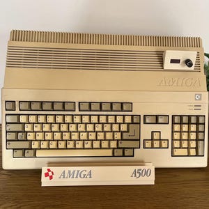 May include: A beige Amiga 500 home computer with a QWERTY keyboard, a numeric keypad, and a top-mounted dial control. A rectangular white sign with the red Amiga logo and the text Amiga A500 is positioned in front of the computer unit.