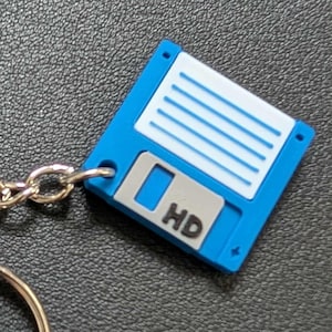 May include: A blue and white floppy disk keyring with a silver chain and a circular key holder. The keyring has the letters "HD" printed on it. The background is a textured black surface.