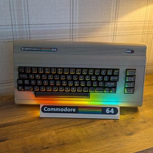 May include: A vintage Commodore 64 computer with a black keyboard and rainbow-colored light. The computer is beige and sits on a wooden surface. A sign below the computer reads "Commodore 64" with a rainbow design.
