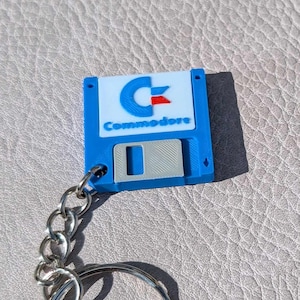 Può includere: Un portachiavi blu a forma di floppy disk Commodore con catena e anello argentati. Il portachiavi presenta il logo Commodore in bianco, blu e rosso. Il floppy disk ha una parte metallica scorrevole argentata.