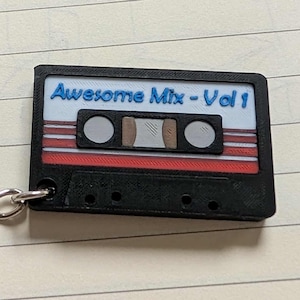 May include: A black cassette tape keychain with the text "Awesome Mix - Vol 1" in blue. The cassette has red and white stripes and silver chain links attached. The keychain is on a lined paper background.