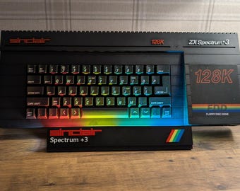 May include: A black Sinclair ZX Spectrum +3 computer with a keyboard and a rainbow-colored stripe. The text "128K" and "Spectrum +3" are visible. The keyboard keys are illuminated with various colors.