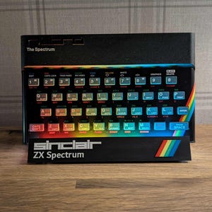 May include: A black Sinclair ZX Spectrum computer with a rainbow stripe design. The keyboard has multiple coloured keys and the text "The Spectrum" is visible. The computer is on a wooden surface.