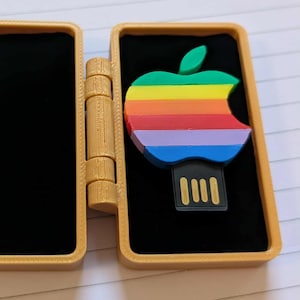 May include: An open, gold-coloured hinged case with a black interior, displaying a rainbow-coloured apple-shaped USB drive. The USB drive has a black connector. The case is made of a textured material.
