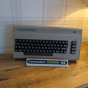 May include: A beige Commodore 64 home computer featuring a dark gray keyboard with black keys, a power indicator light, and a rainbow logo. A white rectangular sign with the text Commodore 64 and a rainbow stripe is positioned in front.