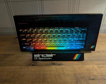 May include: Black Sinclair ZX Spectrum+ computer with a rainbow-colored keyboard. The keyboard keys are illuminated with a gradient of colors. A separate black stand with the Sinclair logo and rainbow stripe is in front.