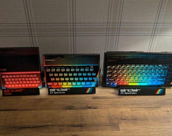 May include: Three vintage Sinclair ZX Spectrum computers are displayed on a wooden surface. The keyboards have rainbow-colored keys and the text "Sinclair ZX Spectrum" is visible. The ZX81 model has red lighting.
