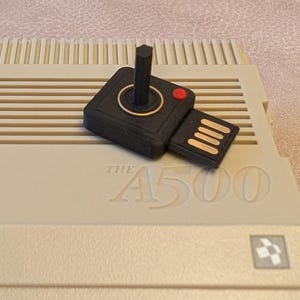 May include: A black USB drive, designed as a retro joystick with a red button and gold detailing, sits on a beige computer. The computer features the embossed text "THE A500". This is a retro gaming accessory.