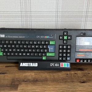 May include: A black Amstrad CPC 464 Colour Personal Computer with a QWERTY keyboard featuring green, red, and blue keys. The unit includes a built-in cassette deck on the right side and a separate black display stand labelled Amstrad CPC 464.