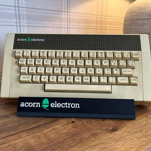 May include: A vintage Acorn Electron computer featuring a beige plastic chassis and a QWERTY keyboard with off-white keys. The top panel has a dark grey grid pattern with the text acorn electron. A black display stand with the logo is in front.