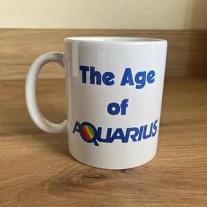 May include: A white ceramic mug with a handle on the left side. The text "The Age of AQUARIUS" is printed in blue on the front. The letter Q in the word AQUARIUS features a small circular icon with horizontal red, yellow, and green stripes.
