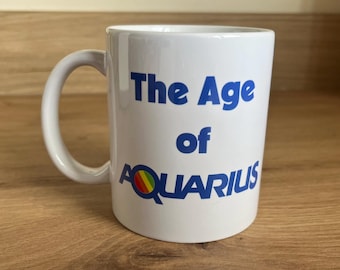 May include: A white ceramic mug with a handle on the left side. The text "The Age of AQUARIUS" is printed in blue on the front. The letter Q in the word AQUARIUS features a small circular icon with horizontal red, yellow, and green stripes.