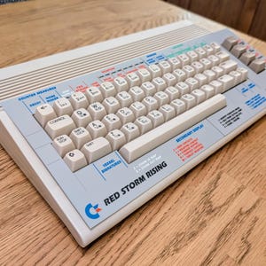 May include: A white computer keyboard with a blue and red logo that says "Red Storm Rising". The keyboard has a variety of keys, including function keys, arrow keys, and number keys. The keyboard is on a wooden surface.