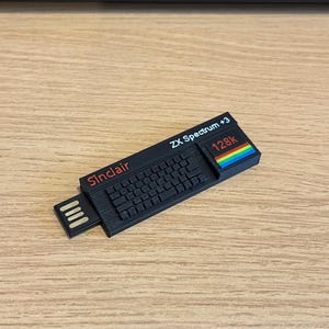 May include: A black USB flash drive shaped like a Sinclair ZX Spectrum +3 computer. The device features a moulded keyboard, the text Sinclair in red, ZX Spectrum +3 in white, and a 128k label with a multi-coloured rainbow stripe graphic.