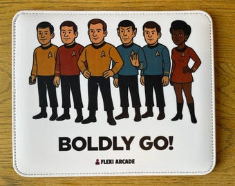May include: A rectangular white mousepad featuring a cartoon illustration of six characters in science fiction uniforms. Below the characters, the text reads BOLDLY GO! and includes the small logo text FLEXI ARCADE at the bottom.