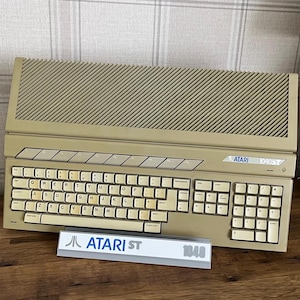 May include: A beige Atari 1040ST home computer featuring a full QWERTY keyboard, a numeric keypad, and a top-mounted ventilation grille. A small rectangular sign in front displays the Atari ST logo and the number 1040 in blue and gray text.