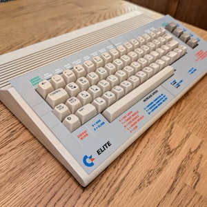 Può includere: Un computer vintage bianco e grigio con una tastiera e uno schermo. Il computer ha un logo blu e bianco che dice "ELITE".