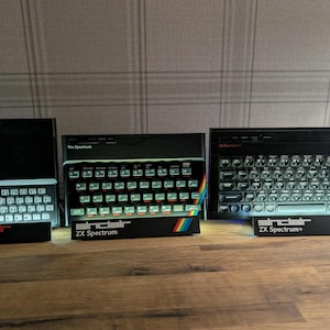 May include: Three black Sinclair computers: ZX81, ZX Spectrum, and ZX Spectrum+. Each has a keyboard and a rainbow stripe. The ZX81 has the text "ZX81" and "Sinclair" in red. The ZX Spectrum has the text "The Spectrum" and "Sinclair ZX Spectrum". The ZX Spectrum+ has the text "ZX Spectrum+" and "Sinclair".