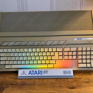 May include: A beige Atari 1040ST computer with a QWERTY keyboard and a diagonal-slotted top vent. The console features the Atari logo and 1040ST text. A white display stand in front reads Atari ST 1040 in blue and grey text, with rainbow lighting.