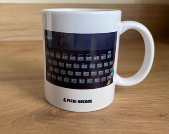 May include: A white ceramic mug featuring a graphic print of a ZX Spectrum computer keyboard with a rainbow stripe on the right side. The text "FLEXI ARCADE" is printed in black below the keyboard image, accompanied by a small red icon.