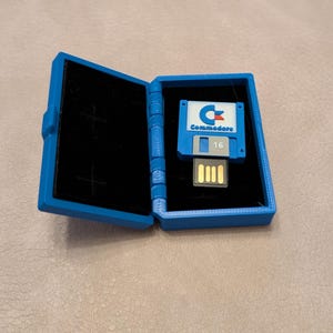 Könnte beinhalten: Ein blaues Plastikgehäuse mit schwarzem Innenraum, geöffnet, um einen blauen und weißen Commodore 16 GB USB-Stick in Form einer Diskette zu zeigen.