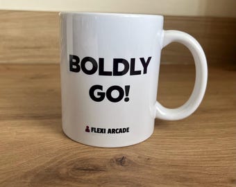 May include: A white ceramic mug featuring the black text BOLDLY GO! printed in a bold sans-serif font. Below the main text, a small red icon of a person is visible next to the black text FLEXI ARCADE. The mug has a standard handle on the right.