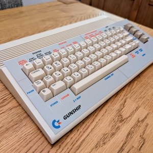 Puede incluir: Un teclado de computadora vintage blanco y azul con la palabra "GUNSHIP" impresa en la parte delantera. El teclado tiene una variedad de teclas, incluidas teclas de función, teclas de flecha y un teclado numérico. El teclado está sobre una superficie de madera.