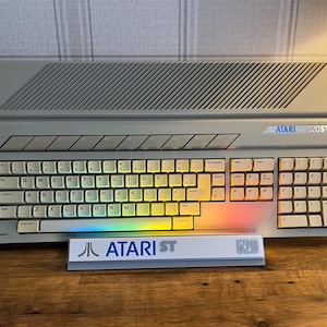 May include: A light gray Atari 520ST computer keyboard with a QWERTY layout, featuring a top ventilation grill and a separate white display stand in front with the Atari logo and the text ATARI ST 520 printed in blue and gray lettering.