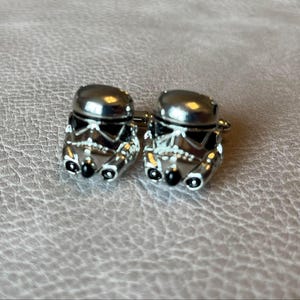 May include: A pair of silver-toned cufflinks featuring a Stormtrooper helmet design. The cufflinks have a detailed, three-dimensional appearance with black accents on a textured background.